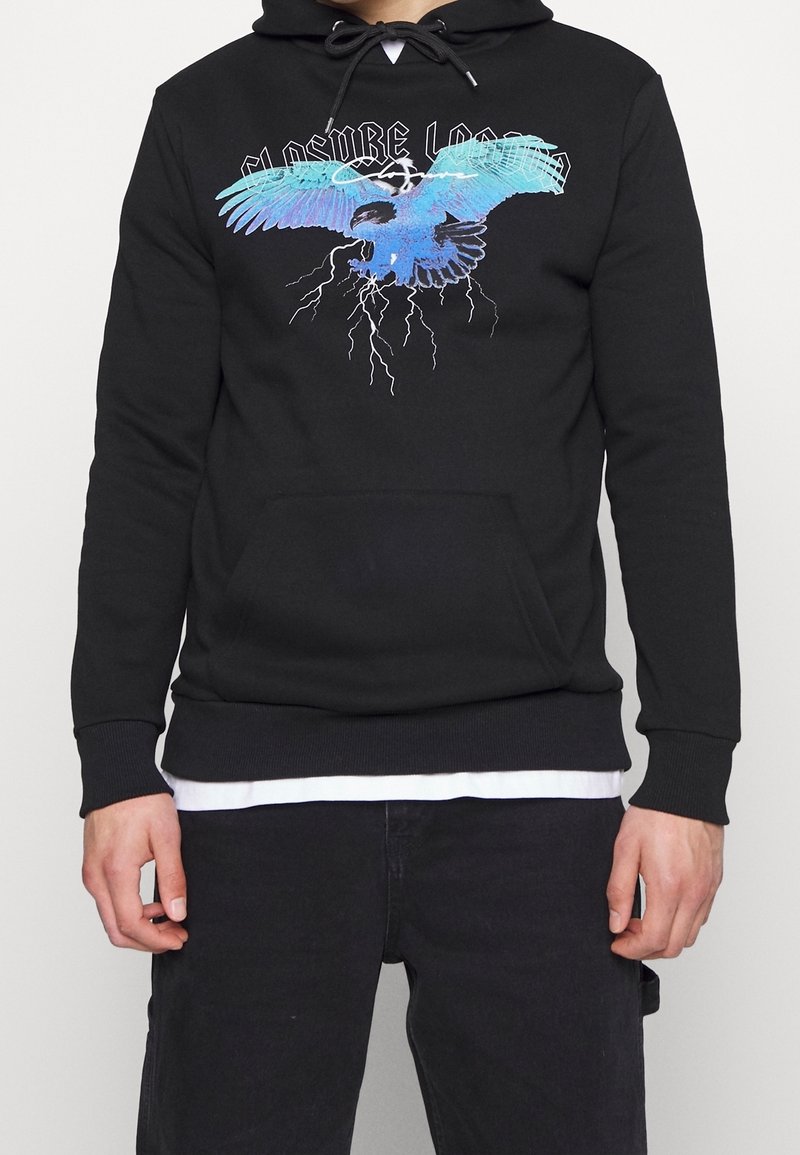 Sweat à capuche noir avec un motif graphique coloré d'un oiseau bleu en plein vol et des éclairs, porté sur une chemise blanche et assorti à un pantalon noir.