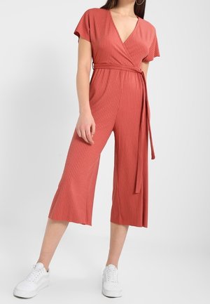 Frau trägt einen rostfarbenen, gerippten Jumpsuit mit kurzen Ärmeln, weitem Bein und gebundenem Taillengürtel, kombiniert mit weißen Sneakers.