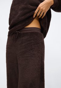 Hnědá fleecová souprava na domácí nošení, která obsahuje měkkou, texturovanou mikinu a odpovídající kalhoty s elastickým pasem a detaily na šňůrku.