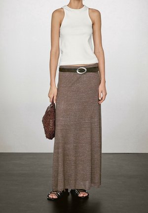 Woman wearing white sleeveless top, brown maxi skirt with belt, black sandals, and holding a brown textured clutch bag.