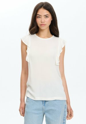 Joven mujer con cabello largo y oscuro que lleva una blusa blanca sin mangas con volantes en los hombros y vaqueros azul claro sobre un fondo blanco.