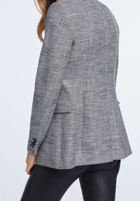 Blazer a scacchi in tessuto bianco e nero, con vestibilità sartoriale, due tasche e polsini con bottone. Texture liscia con un design strutturato.