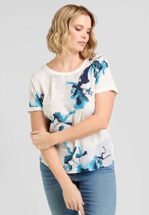 Donna bionda che indossa una camicia bianca a maniche corte con stampa floreale blu e jeans azzurri chiari, in piedi con le braccia incrociate, guarda verso il basso.