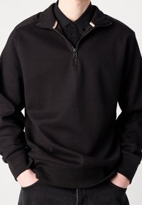 Svart zip-neck sweatshirt med en strukturerad stickad design, med hög krage och ribbade ärmslut, bärs över en svart pikétröja.