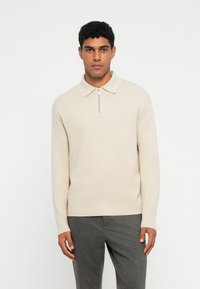 Beige džemperis ar rāvējslēdzēja apkakli, strukturēts audums, pieguļošs design, un garām piedurknēm, kombinēts ar pelēkām biksēm.