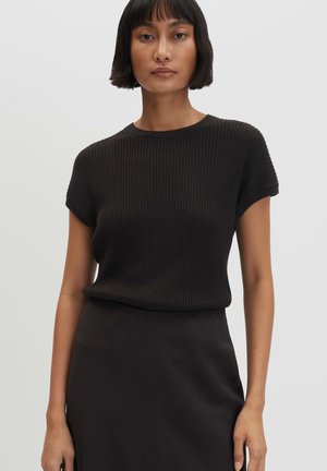 Femme aux cheveux noirs courts portant un haut noir côtelé à manches courtes et une jupe noire, debout devant un fond clair uni.