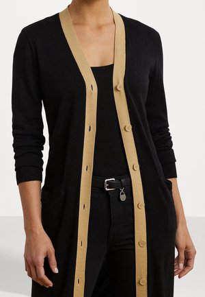 Frau trägt eine schwarze langärmelige Strickjacke mit beigefarbenen Knöpfen über einem schwarzen Oberteil und einem schwarzen Gürtel mit silberner Schnalle.