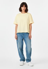 Vrouw met schouderlang donker haar, gekleed in een losse lichtgele T-shirt, ontspannen blauwe spijkerbroek en witte sneakers, staande voor een witte achtergrond.