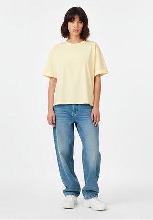 Femme aux cheveux châtains mi-longs portant un t-shirt beige clair ample, un jean bleu décontracté et des baskets blanches, se tenant contre un fond blanc.