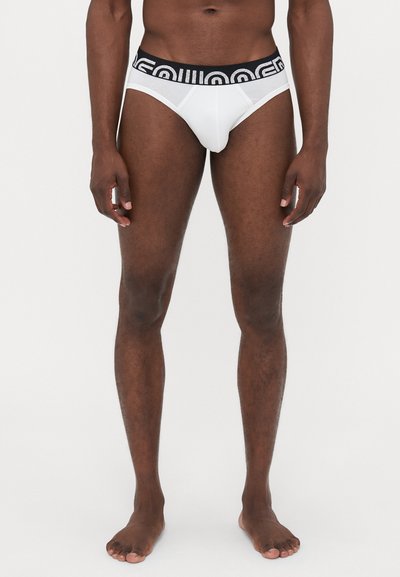 Un slip blanc avec une ceinture élastique noire, arborant un design de logo. Fabriqué en tissu lisse, offrant une silhouette ajustée et une couverture minimale.