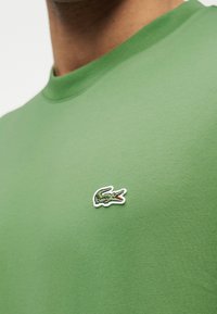 Πράσινο μπλουζάκι με στρογγυλή λαιμόκοψη και μικρό κεντημένο λογότυπο κροκόδειλου στο αριστερό στήθος, που φοριέται από άτομο με μεσαίο τόνο δέρματος.