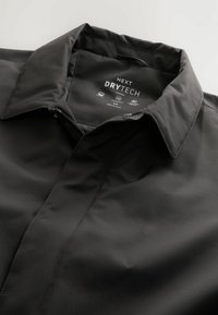 Schwarze Drytech-Jacke mit strukturiertem Kragen und Reißverschluss. Glatte Stoffoberfläche mit matter Optik. Markenlogo innen am Kragen sichtbar.