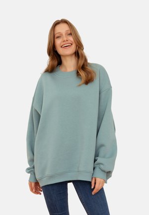 Przesadzony jasnoniebieski sweatshirt uszyty z miękkiego materiału. Posiada okrągły dekolt i opadające ramiona, w połączeniu z dopasowanymi dżinsami.