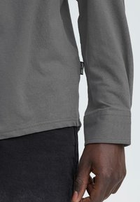 Chemise gris à manches longues avec un col rond, une texture douce et des poignets cousus. Étiquette visible sur la couture latérale. Pantalon foncé en arrière-plan.