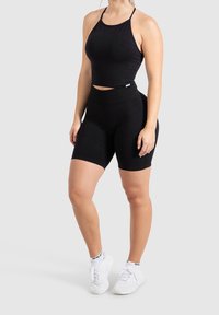 Černý sportovní crop top s tenkými ramínky a odpovídajícími vysokými cyklistickými šortkami z pružného materiálu, doplněný o bílé tenisky.
