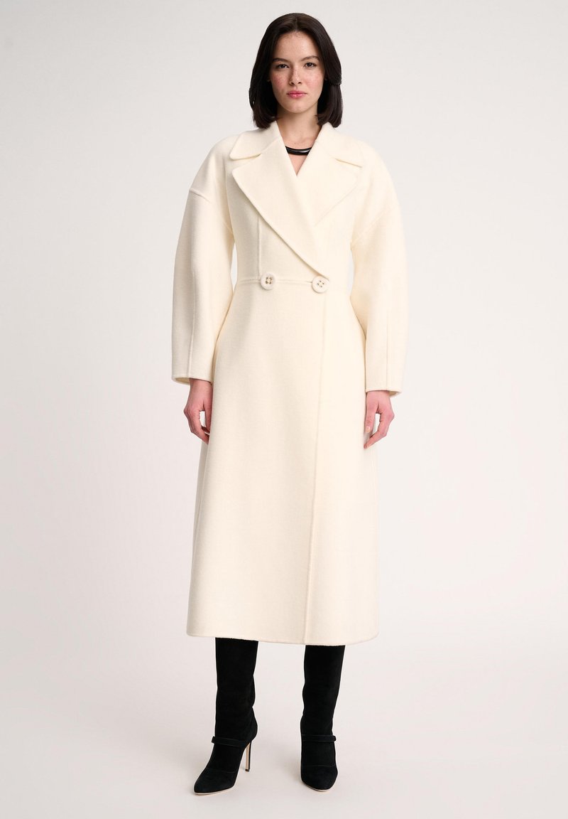 White wool blend coat with a double-breasted design, wide lapels, and two buttons. Paired with black knee-high heeled boots.