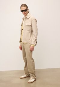 Mees, kes kannab beeži mantlit, kreemjat kardiganit, sobivaid pükse, valgeid treeningjalatseid ja musti prille, seisab tasasel põrandal beeži seina taustal.