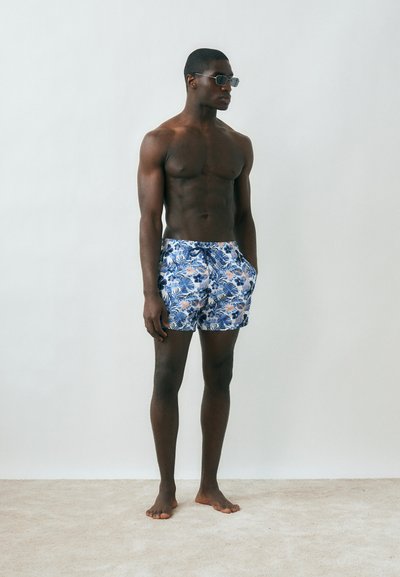 Άνδρας χωρίς πουκάμισο που φορά μπλε και άσπρο μαγιό με λουλούδια και γυαλιά ηλίου, στέκεται ξυπόλητος μπροστά σε απλό ανοιχτό φόντο.