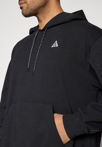 Μαύρο φούτερ με κουκούλα και τριγωνικό λογότυπο στο στήθος, ρυθμιζόμενο κορδόνι με υφή και μπροστινή τσέπη.
