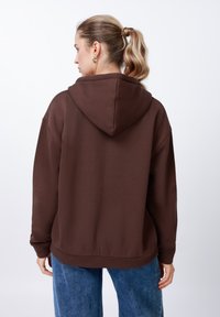 Brūns džemperis ar kapuci, teksturētu virsmu un vaļēju, ērtu piegriezumu. Kombinēts ar gaiši zilām, brīvā piegriezuma džinsu biksēm.