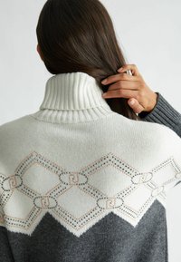 Maglione a collo alto grigio e bianco con colletto a costine. Presenta una cucitura decorativa a motivo diamante e dettagli metallici sottili.