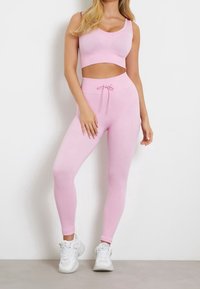 Kobieta w różowej, prążkowanej, bezrękawnikowej bluzce crop top i dopasowanych, wysokich legginsach, w białych sneakersach, stojąca przy białej ścianie.