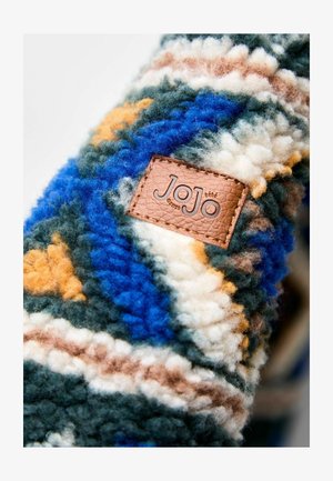 Textil de lana con patrones en azul, verde, naranja y blanco. Presenta un parche de cuero marrón con la marca "JoJo". Superficie gruesa y texturizada.