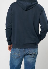 Sudadera navy con un acabado texturizado y capucha con cordón, combinada con vaqueros de denim azul claro y desgastado que presentan diseños curvados en los bolsillos traseros.