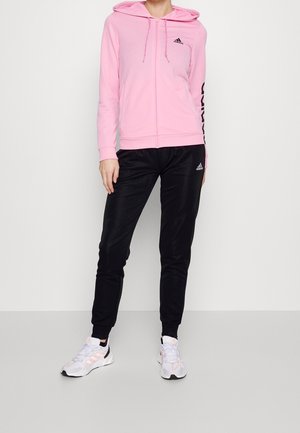Personne portant un sweat à capuche zippé rose d'Adidas, un pantalon de jogging noir Adidas et des baskets blanches Adidas, debout devant un fond clair uni.