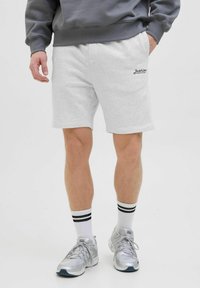 Pantalones cortos de algodón gris claro con bolsillos laterales, presentando un detalle de logo bordado. Combinados con calcetines blancos con rayas negras y zapatillas plateadas.