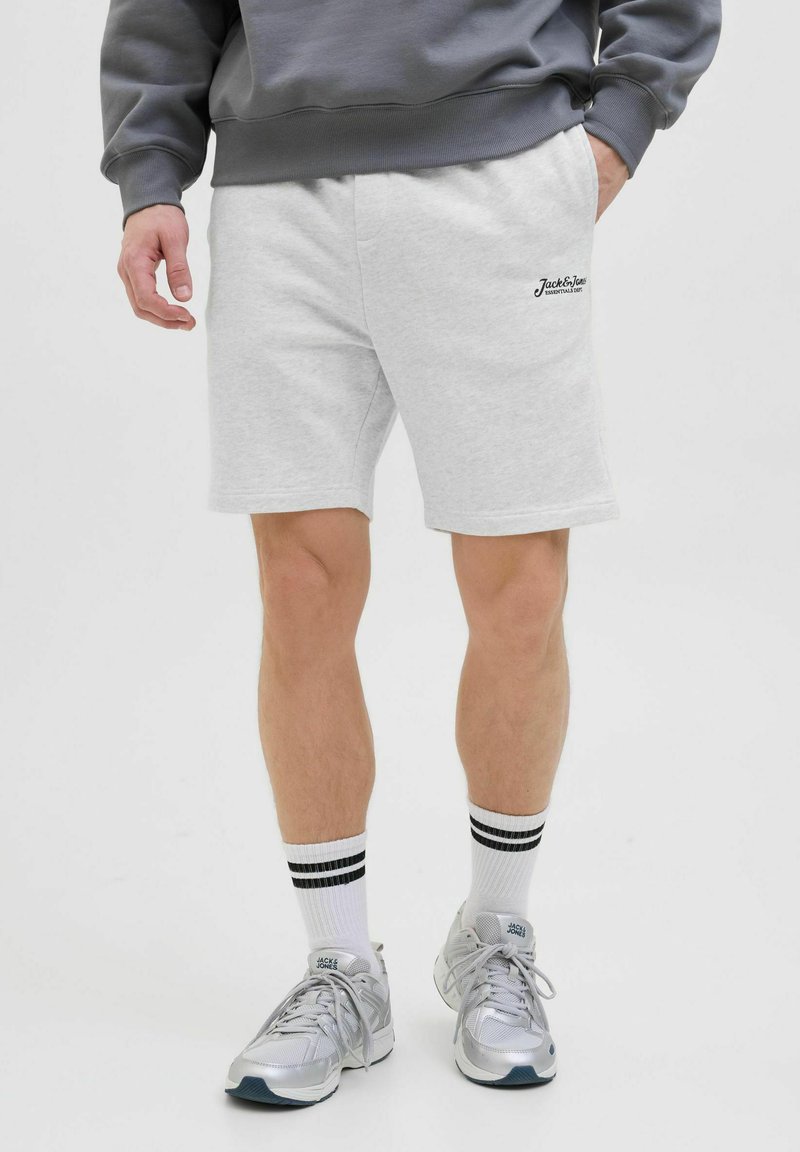 Pantalones cortos de algodón gris claro con bolsillos laterales, presentando un detalle de logo bordado. Combinados con calcetines blancos con rayas negras y zapatillas plateadas.