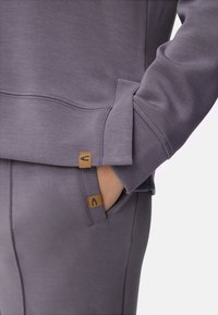 Lichtpaarse sweatshirt met een gladde textuur, voorzien van een kangoeroezak en twee zichtbare merkemblemen bij de naden.