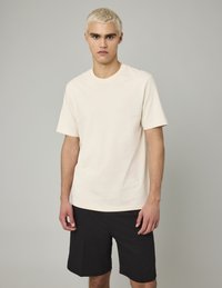 T-shirt en coton beige clair avec un col rond, des manches courtes et une coupe décontractée, associé à un short noir. Texture lisse, sans motifs ni accents.