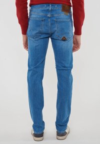 Uomo che indossa jeans blu con tasche posteriori cucite e sneakers bianche, in piedi contro uno sfondo chiaro e neutro.