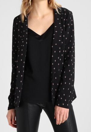 Zwarte blazer met diamantenpatronen in wit en roze, lange mouwen, open voorkant, gecombineerd met een zwarte satijnen cami en leren broek.