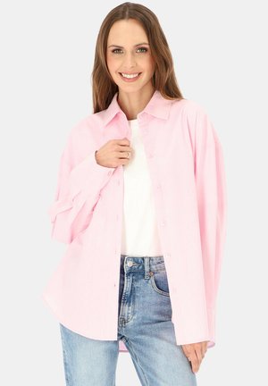 Uśmiechnięta kobieta ubrana w oversize jasnoróżową koszulę zapinaną na guziki, na białym topie i niebieskich dżinsach, stojąca na jednolitym tle.