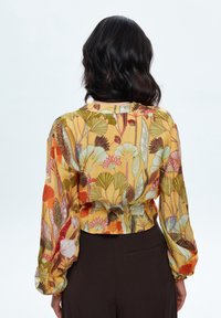 Blusa floreale colorata gialla, con maniche lunghe a sbuffo e design cropped. Presenta intricati motivi di foglie in verde, arancione e marrone.