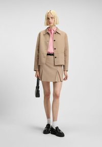 Beige wool-blend jacket with black buttons, pink cotton shirt, beige pleated skirt, black shoes, and a small black handbag.