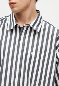 Άνδρας που φοράει πουκάμισο με μαύρες και άσπρες κάθετες ρίγες και κουμπιά, με τσεπάκι στην αριστερή πλευρά, κοιτάζοντας ελαφρώς προς τα αριστερά.