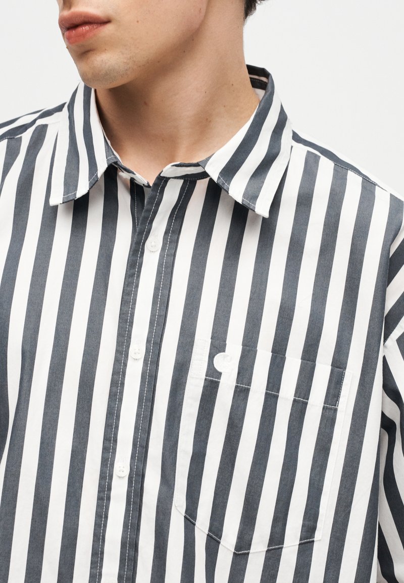 Uomo che indossa una camicia a maniche lunghe a righe verticali bianche e nere con una tasca sul lato sinistro, rivolto leggermente verso sinistra.