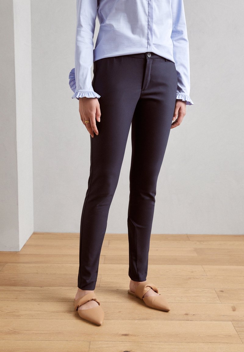 Personne portant un pantalon sombre ajusté, un chemisier bleu clair avec des poignets volantés, des ballerines beiges pointues, debout sur un sol en bois.