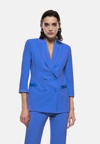 Blazer blu a doppio petto con rever a taglio, maniche a tre quarti e dettagli in satin a contrasto sulle tasche, abbinato a pantaloni coordinati.