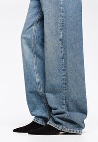 Jeans in denim blu chiaro a gamba ampia con cuciture visibili e orli sfilacciati, abbinati a décolleté nere in camoscio con punta.