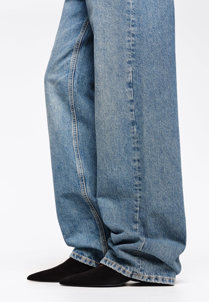 Jeans in denim blu chiaro a gamba ampia con cuciture visibili e orli sfilacciati, abbinati a décolleté nere in camoscio con punta.