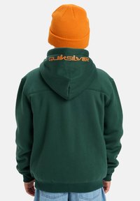 Sudadera de forro polar verde con un logo naranja de "Quiksilver" en la parte posterior. Presenta una capucha con cordón y una textura acogedora. Combinada con un gorro naranja brillante.
