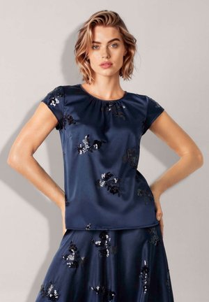 Vrouw met kort blond haar, die een marineblauwe satijnen top en rok draagt met zwarte en zilveren bloemvormige paillettenpatronen, staand met handen in de zij.