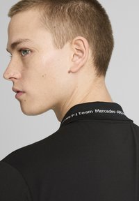 Sort poloshirt med en mærket krave, glat stof og en tætsiddende pasform, vist fra ryggen af en mandlig model.