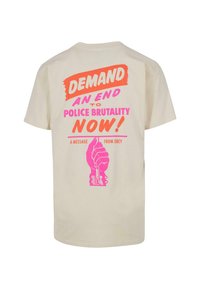 Crème bomulls-t-shirt med djärv grafisk tryck på baksidan. Har orange och rosa text, knytnäve grafik och budskap om polisbrutalitet.