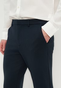 Άνδρας που φοράει ναυτικό μπλε παντελόνι με πλαϊνές τσέπες και λευκό πουκάμισο με κουμπιά, με το χέρι στην τσέπη.