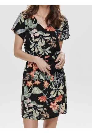 Femme portant une robe noire à manches courtes et col en V, imprimé floral multicolore, ceinture à la taille, et colliers dorés superposés.